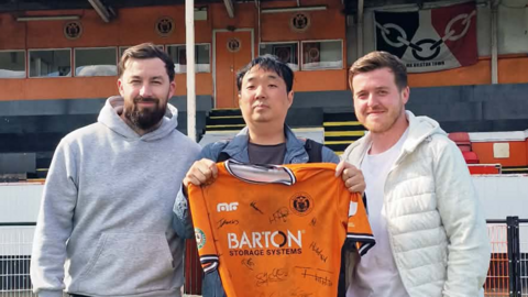 A man with dark hair and a blue jacket holding an orange football shirt between two men with brown hair and white tops with a football stand behind them