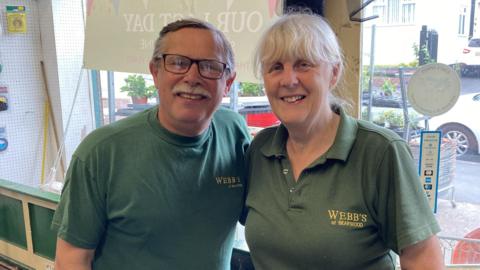 Pauline and Steve Davis, wearing green polo shirts, are stood in the front of their store. There is a window behind them with cars and a street outside.