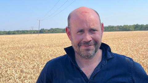 A man with his hands on his hips wearing a denim shirt and he is smiling at the camera. Crops are behind him. 
