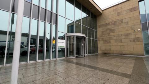 A view of the front of Cumbria House, Cumberland Council's offices on Botchergate, Carlisle. There is a glass front to the building on the left of the picture and an adjoining stone wall straight ahead, with a flagpole in the foreground.