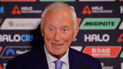 Barry Hearn looks animated during a press conference. He has short grey hair and is wearing a dark suit jacket, white shirt and lilac tie.