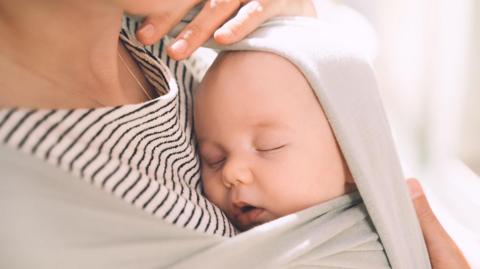 A sleeping baby is being held against her mother who is wearing a stripey brown top and has the baby swaddled against her chest 