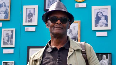 Dr Vanley Burke posing infront of a gallery of his photos, wearing a black bowler hat, sunglasses, a black t shirt and a green jacket