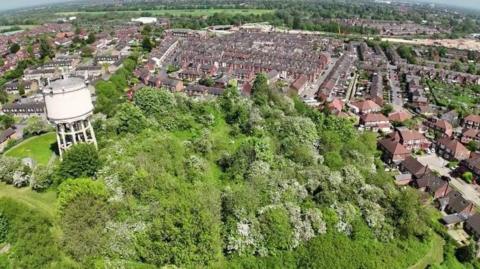 A drone shot showing a mound of grass and greenery with a water tower in the foreground, with streets of houses behind.