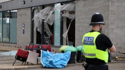 A view of damage to Holiday Inn Express hotel after rioters attacked the building in Rotherham, with chairs scattered around outside and glass from the windows smashed on to the ground. A police officer also folds his arms and looks into the distance next to the damage. 