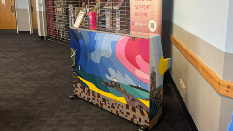 The painted piano on display at the terminal. The piano is painted blue, pink and yellow and depicts a Guernsey landscape. The piano is on display in front of a shop inside the terminal. A large pink banner is on display behind the piano.