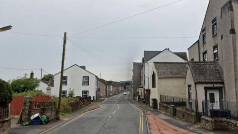 The A5086 Main Street in Cleator: terraced houses with slate roofs line the road on either side; there is a white car is in the distance.