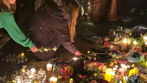 Two women are seen placing flowers at a vigil where there are many candle lit, flowers on the ground and balloons. It is dark and there are fairy lights seen on a low wall in the background