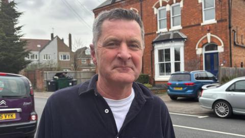 A man in a white t-shirt and navy shirt stands on a residential street on which are several parked cars and looks at the camera unsmilingly 