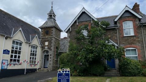  The outside of a school. The building on the left is white with large windows and bunting. The building in front is made of a brown brick and has a green bush growing on it. There are grey clouds in the sky. 