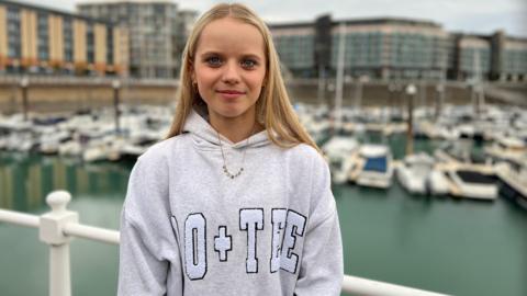 Sofia smiles at the camera. She is wearing a grey hoodie with a necklace. Her blonde is beyond her shoulders. Behind her, there are boats in a marina with buildings further in the background.