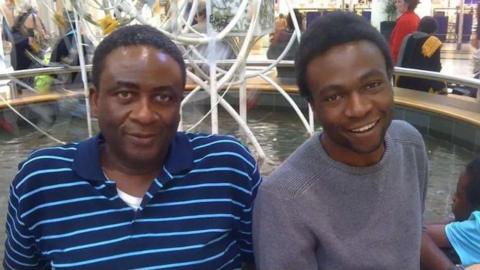 Two men sit side-by-side smiling at the camera. The man on the left is wearing a blue stripy polo shirt while the man on the right is wearing a grey jumper