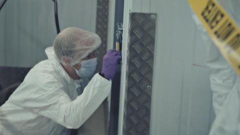 A forensic officer wearing a white hazard outfit and hairnet, purple rubber gloves and a clinical face mask, kneels down next to a storage locker. They are gripping a padlock that is connected to a metal bolt. To one side is yellow "Do not enter" police tape.