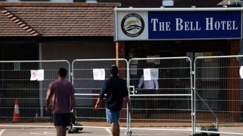 Two men walking away from the picture with only their backs visible towards the bell hotel which is surrounded by metal wire fencing.