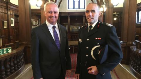 Staffordshire commissioner Ben Adams with fire chief Rob Barber. They are standing in a grand room. Mr Adams is wearing a smart suit with a purple tie and the fire officer is wearing his uniform and holding a hat. 