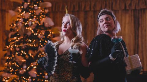 Two women in 1920s evening outfits stand in front of a Christmas tree lit with fairy lights. Everything in the decor and their clothes is themed gold and black.