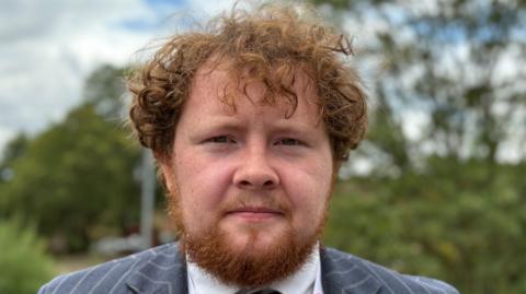 Taylor Cowley-Coulton with long curly brown hair and beard, wearing a blue and white pin-striped jacket and blue and black tie with a lanyard. There is a blurred exterior scene of trees behind him