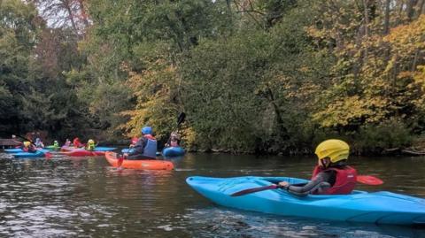Young children in colourful kayaks on a stretch of water with trees on the far bank.