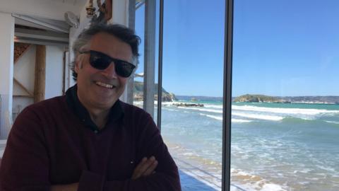 Nicolas Gerasimidis, smiling and wearing sunglasses, sitting by a window of a building by a beach with the sun shining