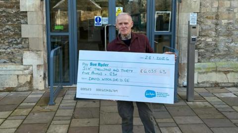 David, a man, with short white hair, wearing a burgundy jumper and grey trousers, holding a large, white cheque in his hand.