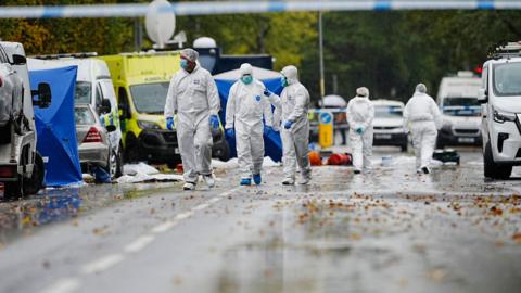Forensics search the streets surrounding the synagogue. Three officers wear white overalls, blue gloves and face masks