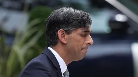 Rishi Sunak facing right, looking quite glum, he is wearing a blue suit with a white shirt and tie