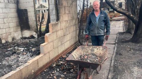 A person stands beside a wheelbarrow filled with rubble next to the fire-damaged remains of a building. Charred walls, burnt trees and ash-covered ground surround the site, while neighbouring homes can be seen in the background.