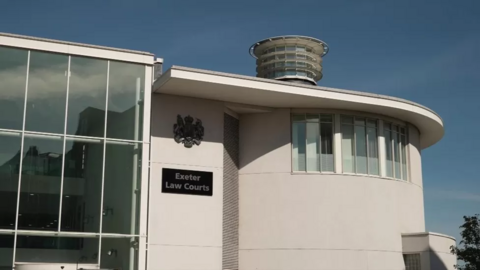 The exterior of the court building which has a sign reading Exeter Law Courts with a heraldic shield above it. Part of the building is floor to roof glass and the other section is curved with a row of windows at the top. On the roof is a short tower with windows all around it reminiscent of a lighthouse. 