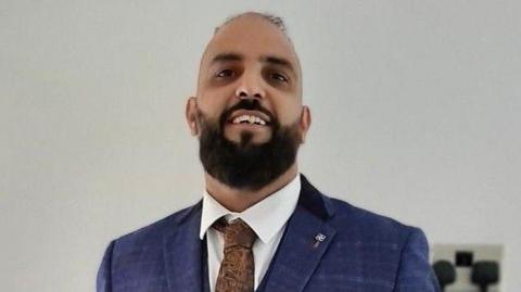 A man stands smiling wearing a blue suit, white shirt and brown tie. He has a short dark beard.