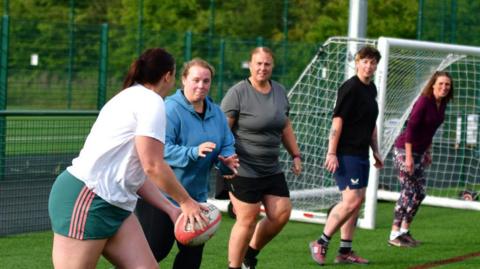 A line of five women playing rugby. To the left, the player closest to the camera is about to pass the ball. The second player is waiting to catch it. The women are wearing shorts and t-shirts of various styles and colours.