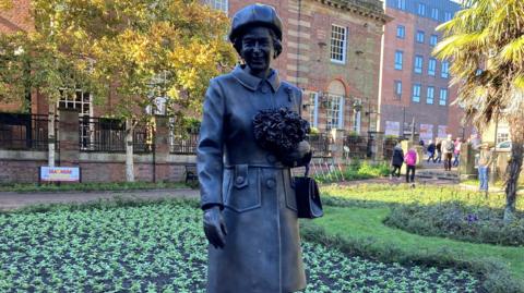 A bronze-cast statue of Queen Elizabeth II - the Queen is wearing a hat and smiling as she holds a bouquet of flowers and a handbag. 