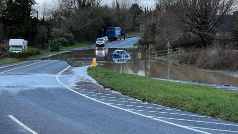 A blue Mini car floats in floodwater on a main road through the Cotswolds. Out of the water, on the road behind, there is a police van and a lorry, and a motorhome can be seen on a grass verge. 