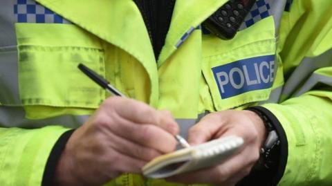 A police officer is taking down notes with a pen and a notepad. He is wearing a fluorescent green jacket with a logo on it that says please in grey writing.