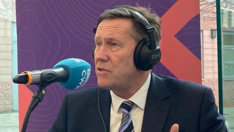 A man wearing a suit sits at desk, speaking into a microphone. Purple boards with the BBC Radio CWR logo and text can be seen behind him.