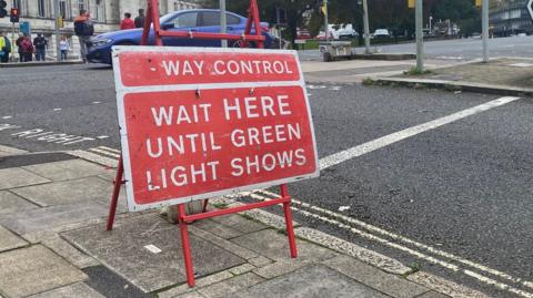 Red temporary traffic sign at junction which reads: "Wait here until green light shows."