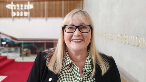 Karen Kilgour smiling at the camera. She has medium-length straight blonde hair with a fringe. She is wearing black-rimmed glasses, a green-and-white patterned shirt and a black suit jacket. 