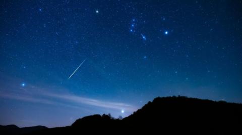 A light blue night sky filled with meteor showers and stars, with the silhouette of a hill underneath it