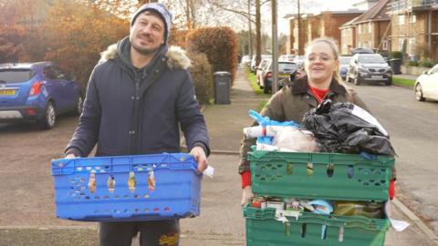 A man and woman carrying crates of food along a residential street. The man has a winter coat on with a fluffy hood and a blue beanie hat. He is carrying one blue crate. The woman has blonde hair and glasses. She is carrying two heavily laden green crates.