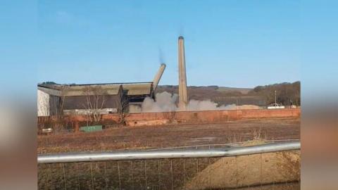 A chimney collapses on an industrial site with billows of dust around it.