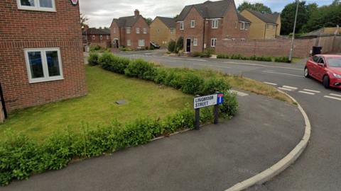 A newbuild housing estate. A red car turns a corner into Lingmoor Street which is identifiable via a street sign.