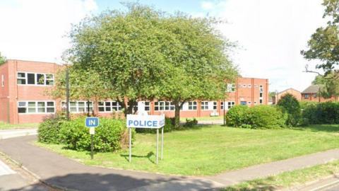 The outside of a police station. It is made of light red bricks. Outside is a large front lawn and trees, and a white sign with the word 'police' in blue capital letters.