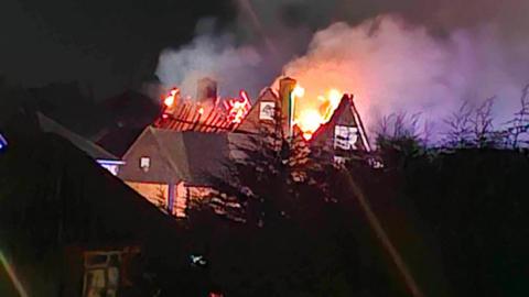 A building on fire - the roof has gone and flames can be seen coming from the top of the building. It is dark outside.  