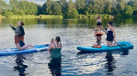 Three women are paddleboarding on a lake. One has red hair, brown shorts and a stripped top and is kneeling whilst laughing. One is also laughing dressed in a navy swimming costume with white trim. The other is paddling whilst standing in a white bathing costume and black shorts. Sh has a small dog standing on the front of her paddleboard. Another lady is paddling up to her waist dressed in a green swimming costume whilst holding a baby in her arms.