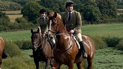 In the foreground are three horses being ridden by hunters wearing tweed jackets, beige jodhpurs, and long black riding boots. The horses are various shades of brown and chestnut - there are more riders and horses in the background. They're riding along a ridge with the Wrekin clearly seen in the background - the weather is sunny and the grass is bright green.