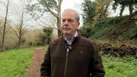 Paul Masterton is wearing a dark green and brown fleece and a light blue and white striped shirt underneath. He has grey hair and is stood in a wooded area. 