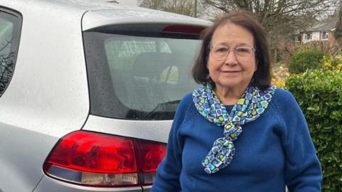 Marbelia Davison pictured outside her home, beside her silver car. Marbelia is wearing a blue jumper, and patterned neckerchief. She has shoulder length hair and is wearing glasses.