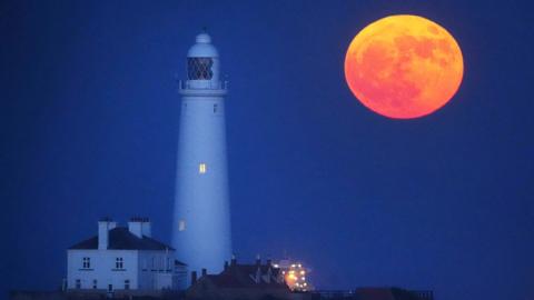 A red-orange full Moon hangs low in the night sky next to a lighthouse