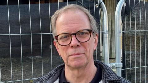 A head shot of Rob Frankson. He is wearing glasses and a black and grey dogtooth patterned coat over a black top. He is outside and silver railings can be seen behind him. 
