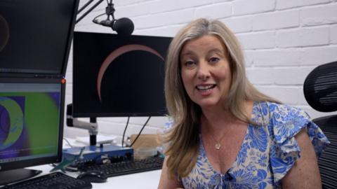 Weather presenter Sara speaks to the camera, with monitors behind her showing the eclipse.