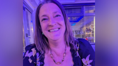 Karina Haselgrove-Spurin is seen from her shoulders up sitting by a window. She is wearing a top with a flour print and is smiling at the camera.